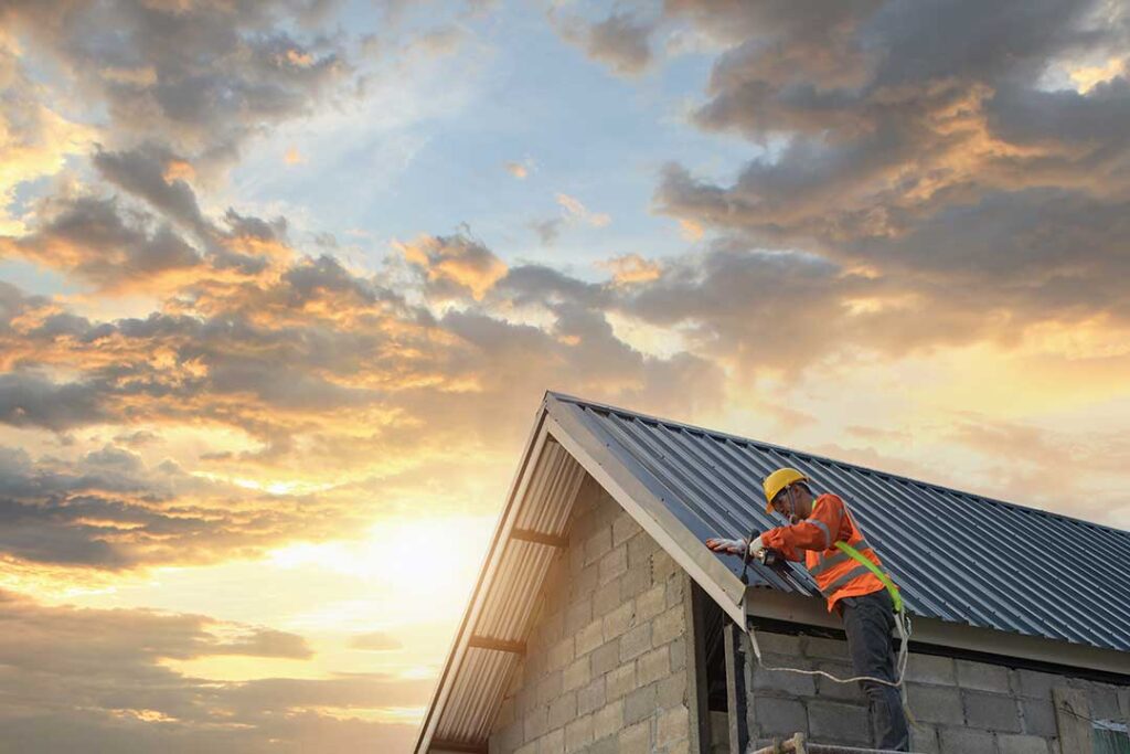 Person clearing leaves from a residential gutter beside a shingled roof with a ladder and bucket.
